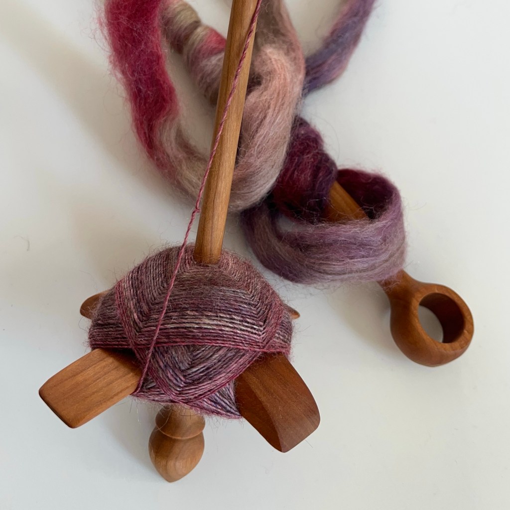 Cherry wood cross-bar spindle loaded with a single of variegated lilac yarn is resting on a white surface. A ring distaff is loaded with unspun fibre and rests behind spindle.